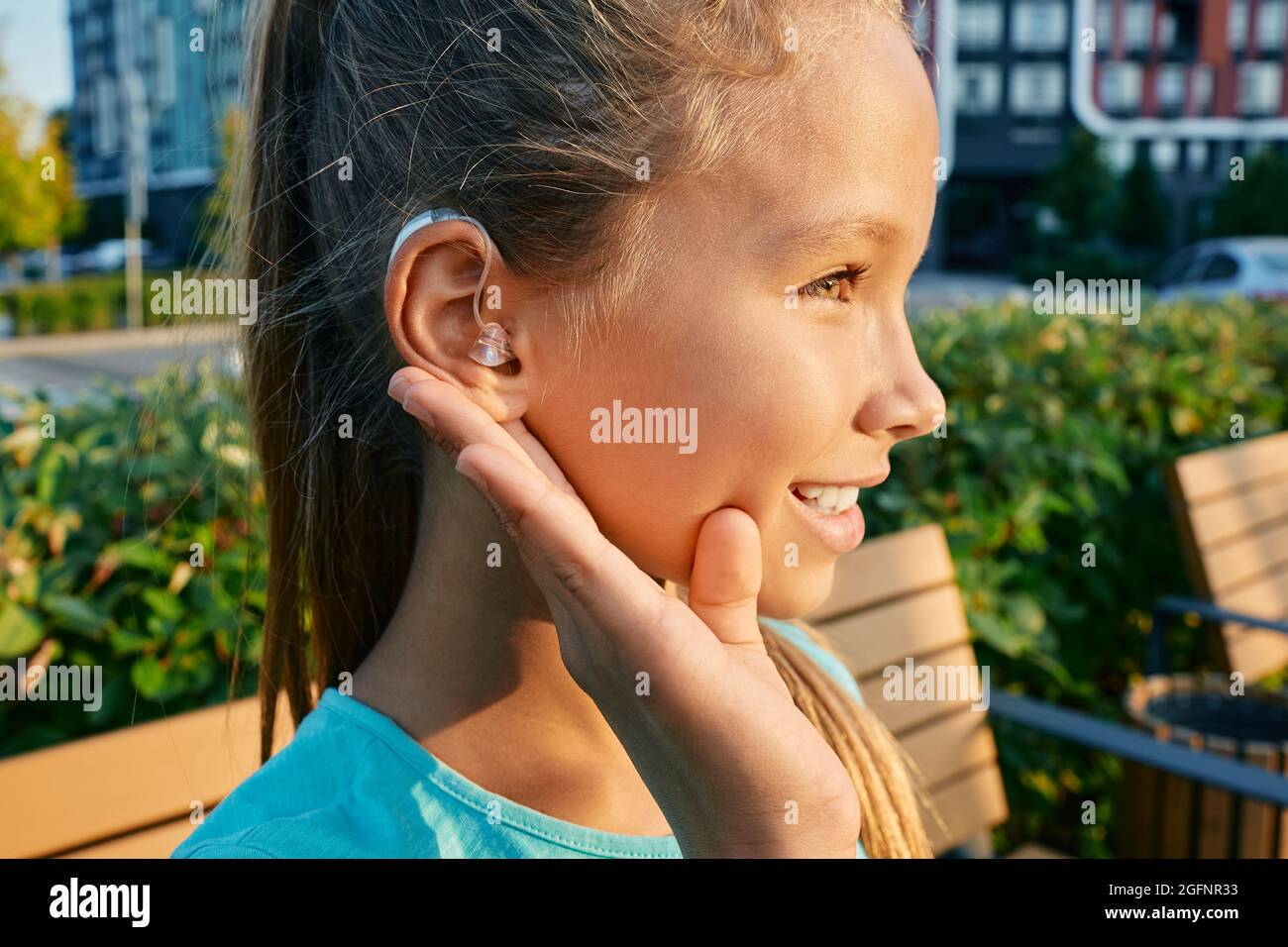 Lächelndes weibliches Kind mit einem Hörgerät hinter dem Ohr hält die Hand nahe am Ohr, um im Freien zu hören und ein Leben lang zu leben Stockfoto