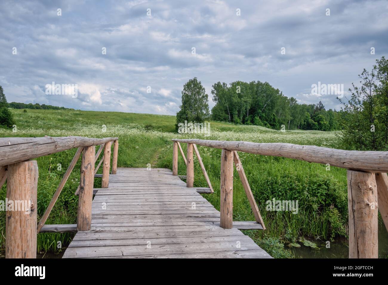 Malerische Landschaft mit einer Holzbrücke über einen kleinen Fluss und Bäumen im Hintergrund. Ländliche Szene gegen den bewölkten Himmel mit Wolken. Das Feld wi Stockfoto