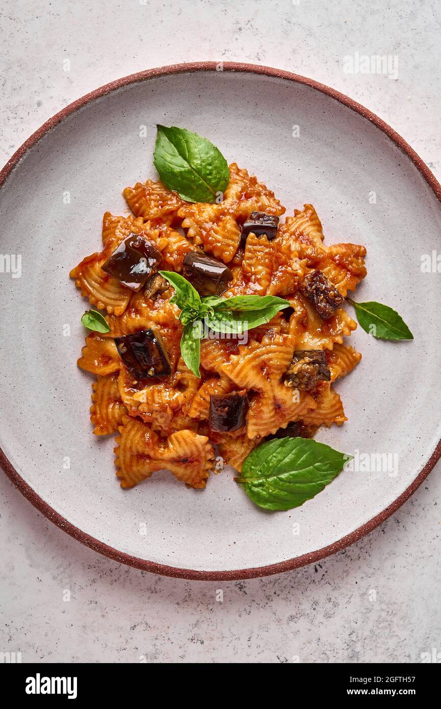 Pasta Farfalle mit Auberginen, Tomatensauce, Käse und Basilikum auf dem Teller auf einem grauen strukturierten Hintergrund. Vertikale Ausrichtung Stockfoto