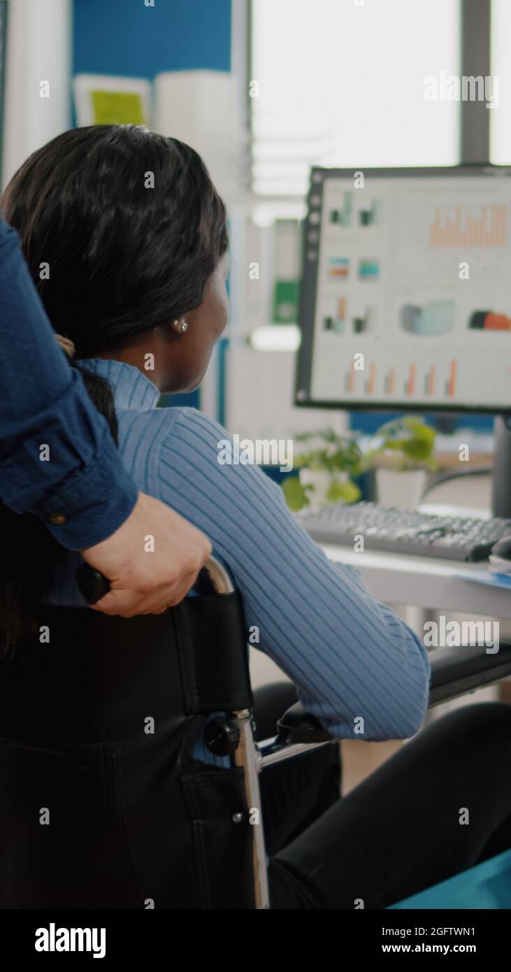 Ein Geschäftsmann hilft seinem behinderten schwarzen Kollegen, der in das Firmenbüro eintritt und den Rollstuhl in die Nähe des Schreibtisches schiebt. Afrikanisch behinderte gelähmte Frau begrüßt Kollegen, die bei einem Finanzprojekt arbeiten Stockfoto