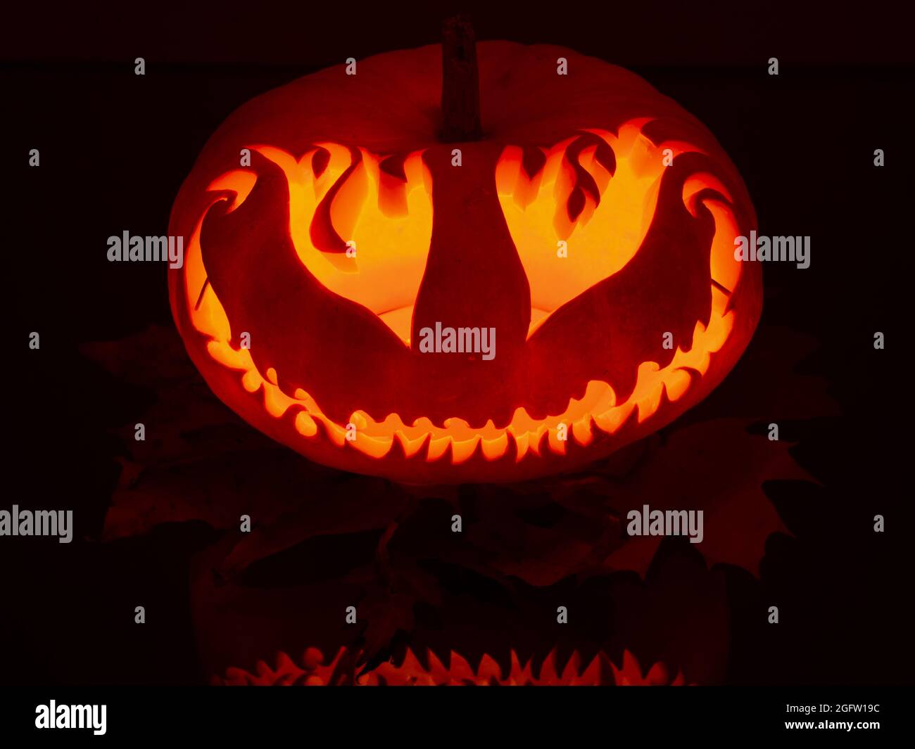 Beleuchteter, lächelnder, großer, handgemachter Kürbis mit Augen wie Feuerflammen auf Ahornblättern in der dunklen halloween-Nacht Stockfoto