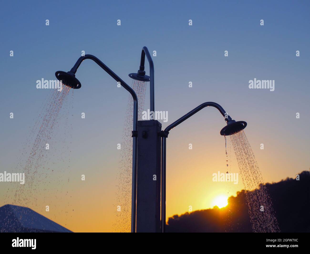 Wasserstrahlen, die aus der dreifachen Stranddusche gegen die untergehende Sonne strömen Stockfoto