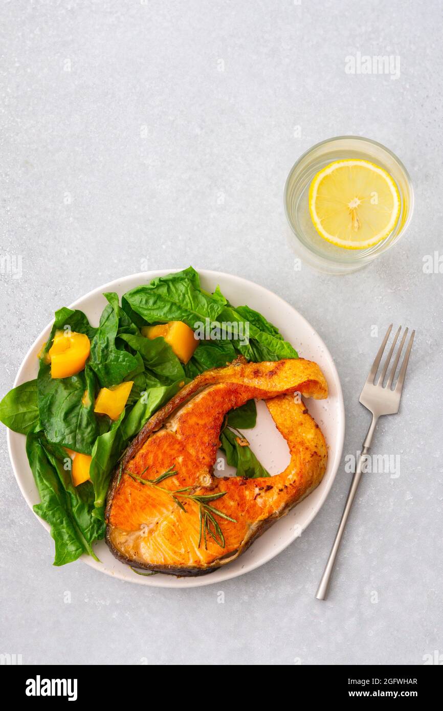 Gebratenes Lachssteak mit Spinat und gelben Tomaten auf einem Teller auf grauem Grund, ein Glas Wasser mit Zitrone, ein herzhaftes Mittagessen mit gesundem rotem Fisch Stockfoto
