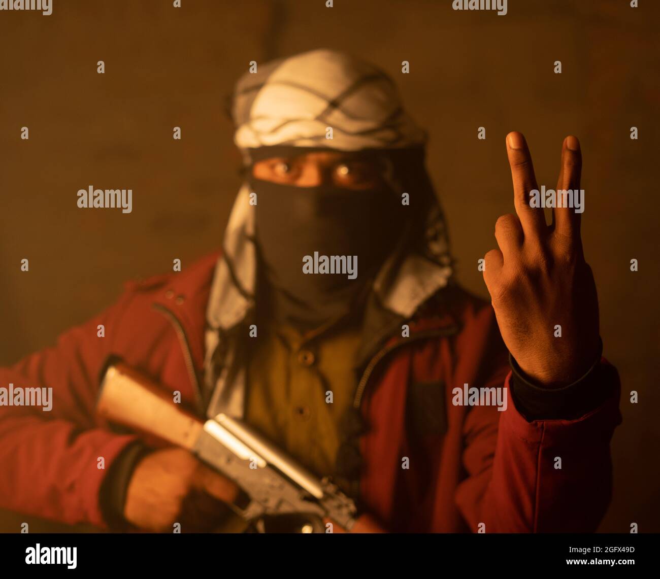 Glücklicher islamischer Militant oder Soldat mit Gesichtsabdeckung, die V-Zeichen oder Siegesgeste auf Kriegsführung mit Waffe in der Hand zeigt Stockfoto