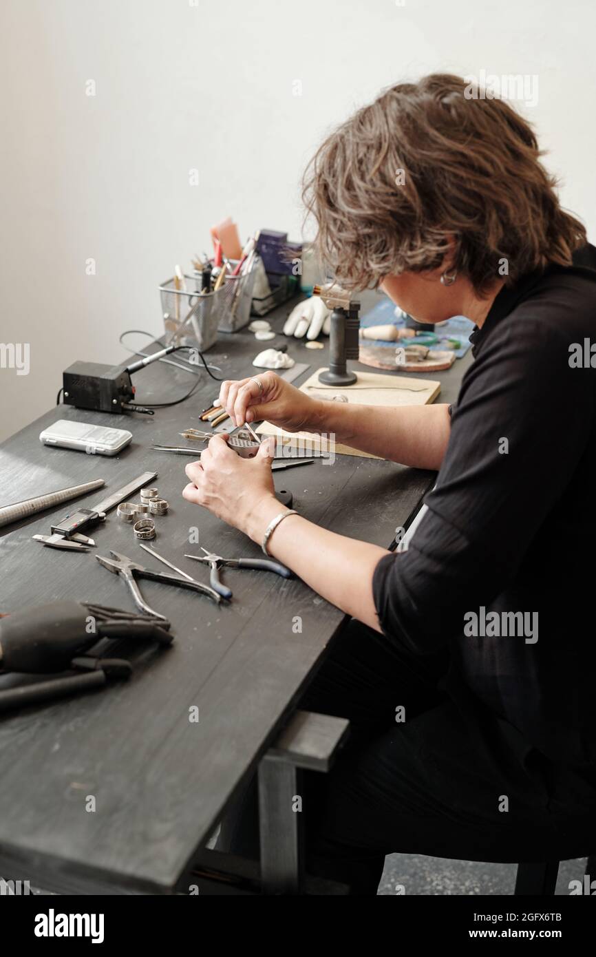 Weibliche Juwelierin sitzt am dunklen Tisch mit Arbeitswerkzeug und macht einen neuen Ring für individuelle Aufträge Stockfoto