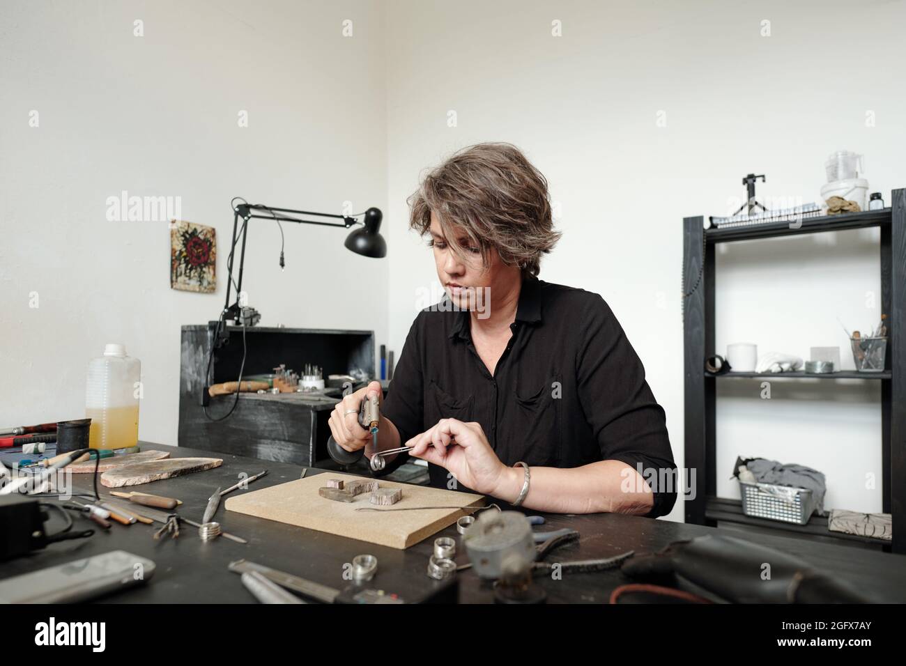 Beschäftigte Frau mittleren Alters im schwarzen Hemd, die in der Werkstatt am Schreibtisch sitzt und Schmuck lötet, während sie die Arbeit beendet Stockfoto