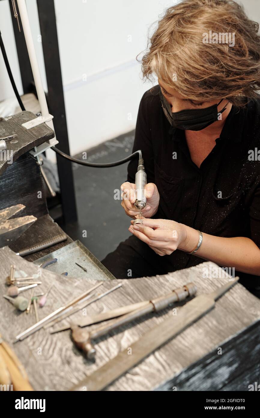 Zusammengezentrierte weibliche Juwelierin in Maske sitzt am Arbeitsplatz und mit abrasiven Düse bei der Erstellung Design des Rings Stockfoto