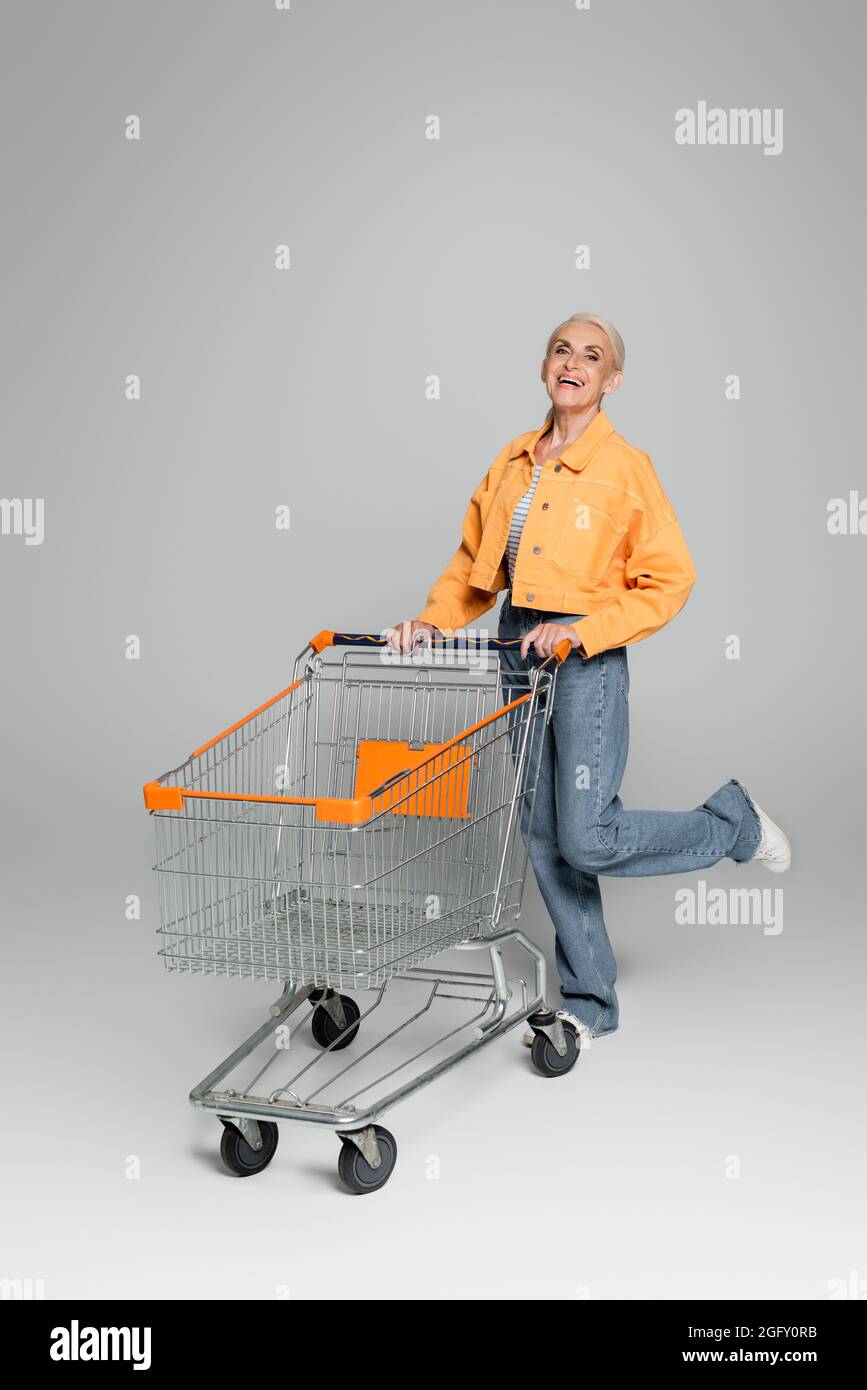 Fröhliche ältere Frau in stilvoller Kleidung, die auf einem Bein in der Nähe des Einkaufswagens auf grau steht Stockfoto