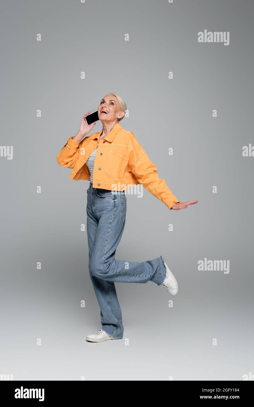Aufgeregte und stilvolle ältere Frau, die auf dem Smartphone spricht, während sie auf einem Bein auf Grau steht Stockfoto