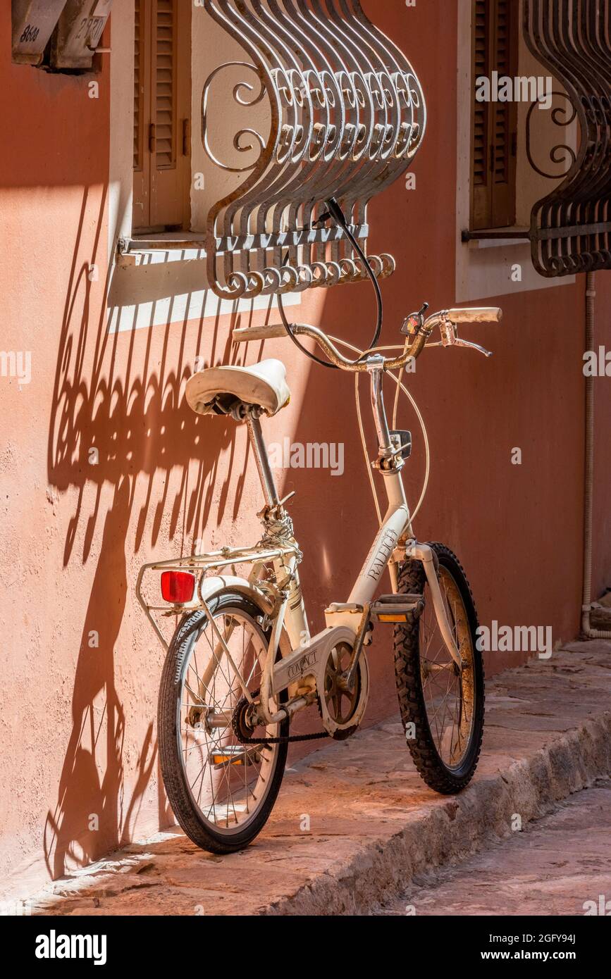 Faltfahrrad, das einen Schatten außerhalb eines griechischen Hauses wirft, typisch griechisch, typisch griechisch, Fahrrad am Fenster lehnend, Fahrrad an einer Wand lehnend. Stockfoto