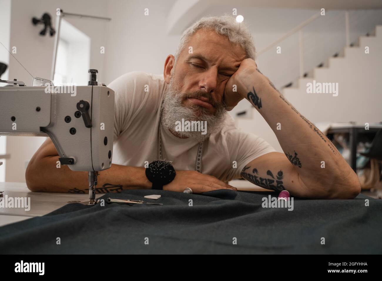 Müder Schneider sitzt in der Nähe von Stoff und Nähmaschine Stockfoto