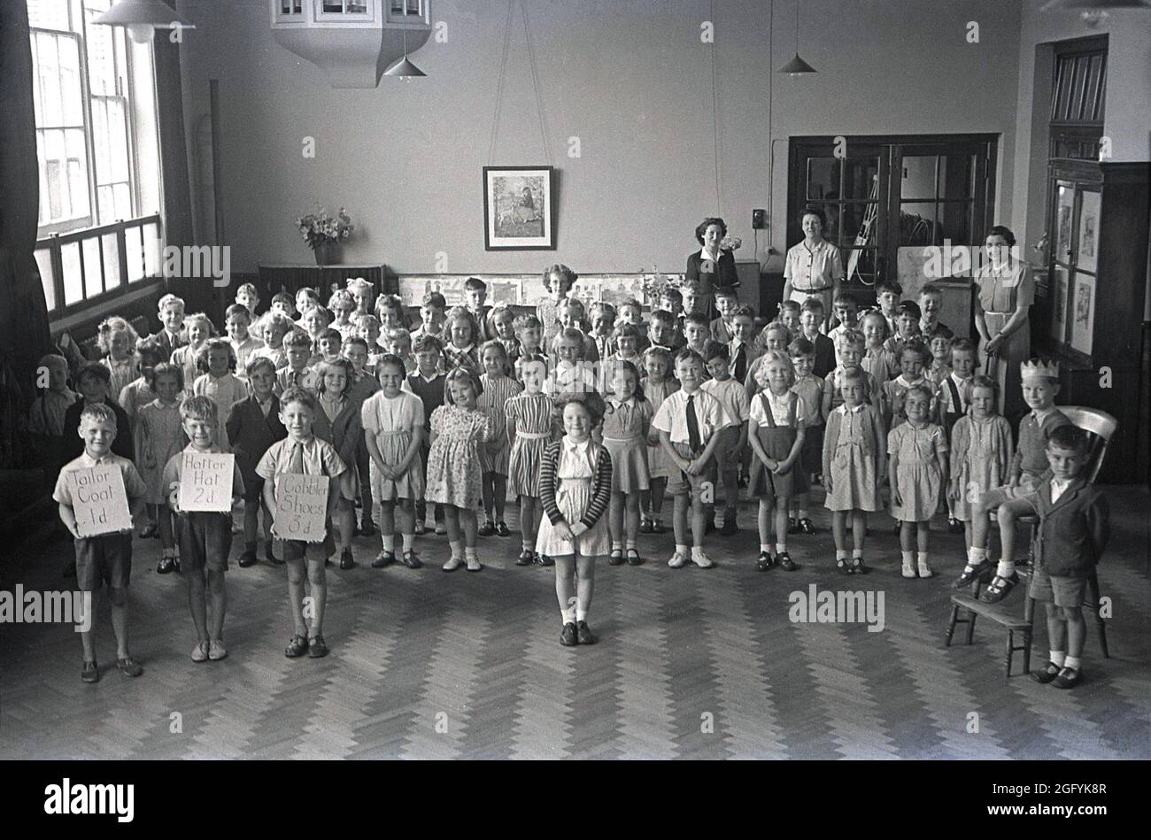 1955, historische Schulversammlung an einer Grundschule, Schüler, die für ein Gruppenbild stehen, mit drei jungen Jungen an der Vorderseite der Gruppe, die Karten hochhalten und sagen: „Tailor Coat 1d“ und Hatter hat 2d“ und „Cobble Shoes 3d“, England, Großbritannien. Stockfoto