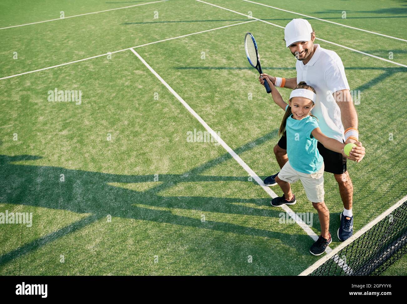 Positive kleine Mädchen beim Lernen, Tennis mit männlichen Trainer auf Outdoor-Rasenplatz zu spielen. Einzellehrer für ein Kind zum Tennistraining Stockfoto