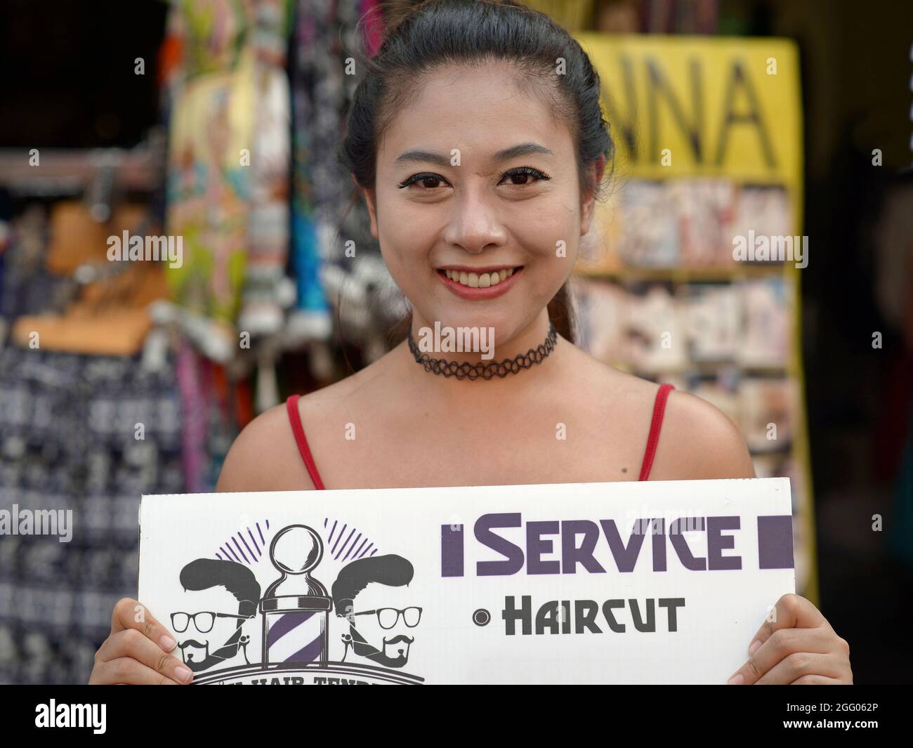 Die fröhliche junge, schöne thailändische Friseurin wirbt auf einem handgehaltenen Schild für ihren Friseurservice und lächelt für den Betrachter. Stockfoto