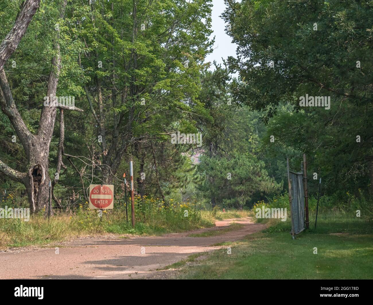 Leere, staubige Schmutzeinfahrt mit "nicht betreten" Schild und Zaun führt durch ein offenes Tor in einen stark bewaldeten Wald. Stockfoto