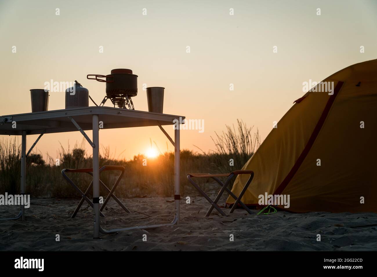 Blick auf den Sonnenuntergang auf eine faltbare Campinggasfeueranlage mit einem Metallglas und einem Topf auf einem Tisch. Wilder Naturtourismus mit Zelt, aktives Lifestyle-Konzept Stockfoto