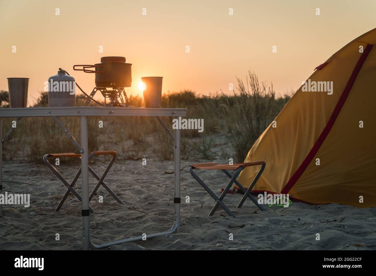 Blick auf den Sonnenuntergang auf eine faltbare Campinggasfeueranlage mit einem Metallglas und einem Topf auf einem Tisch. Wilder Naturtourismus mit Zelt, aktives Lifestyle-Konzept Stockfoto