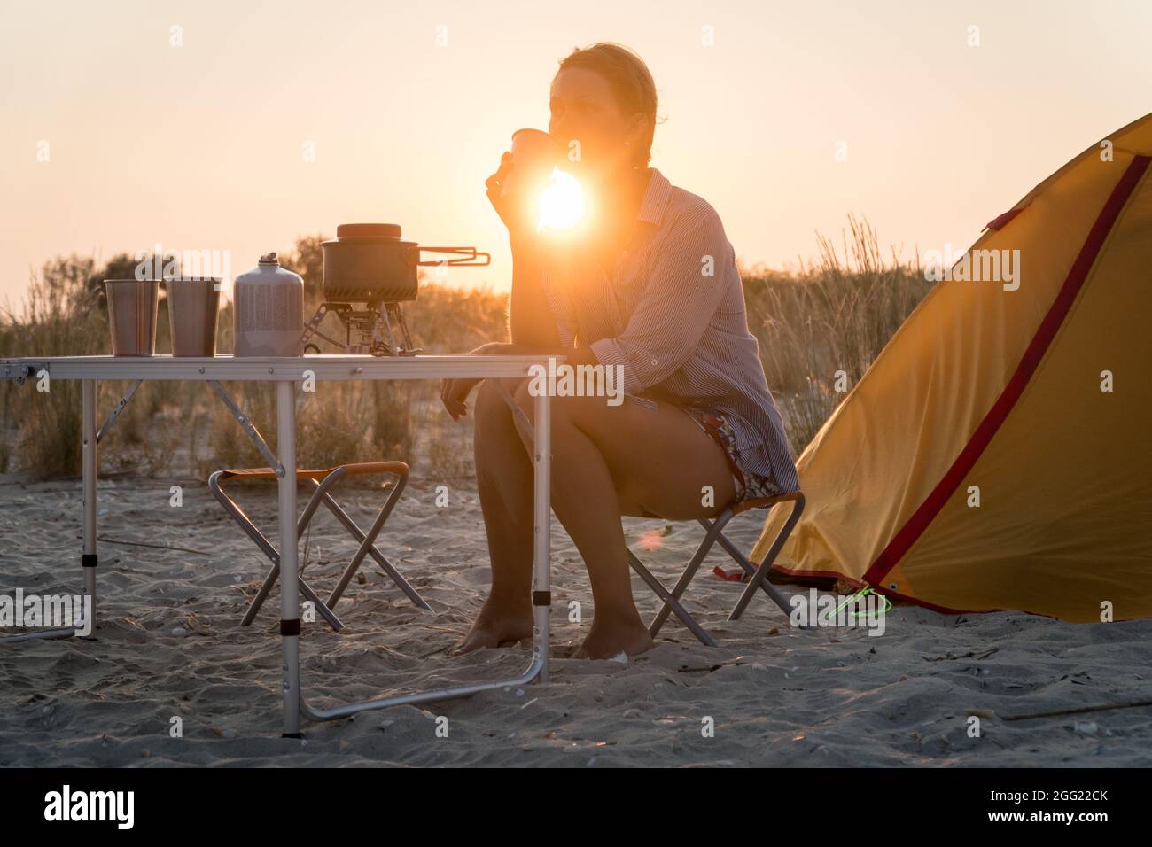 Sonnenuntergang szenische Ansicht der Frau, die Metallglas hält. Faltbares Campinggas-Feuersystem mit einem Topf auf einem Tisch. Wilder Naturtourismus mit Zelt, aktiv lebend Stockfoto