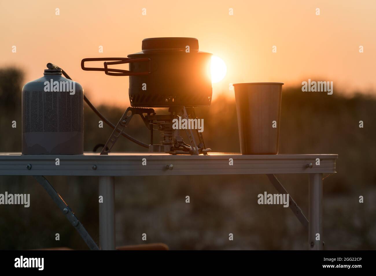 Blick auf den Sonnenuntergang auf eine faltbare Campinggasfeueranlage mit einem Metallglas und einem Topf auf einem Tisch. Wilder Naturtourismus und aktives Lifestyle-Konzept Stockfoto