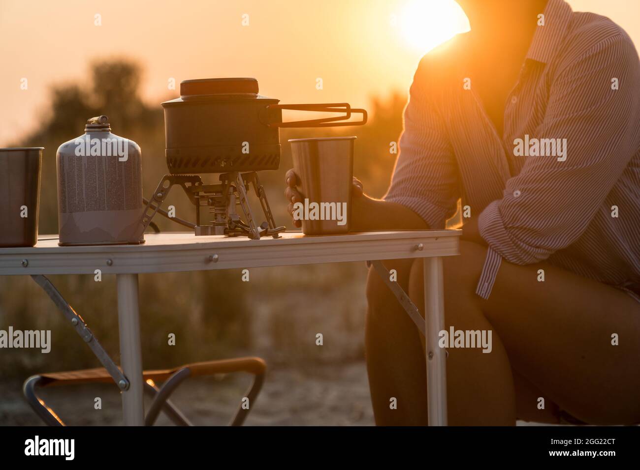 Sonnenuntergang szenische Ansicht der Frau, die Metallglas hält. Faltbares Campinggas-Feuersystem mit einem Topf auf einem Tisch. Wild Nature Resort und Active Lifestyle Konz Stockfoto