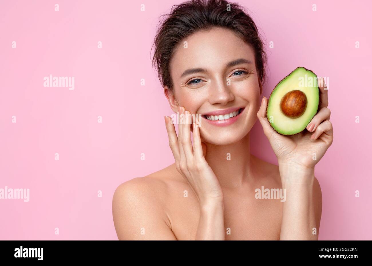 Lachende junge Frau mit einer halben Avocado. Foto von attraktiven Frau mit perfektem Make-up auf rosa Hintergrund. Beauty & Hautpflege Konzept Stockfoto