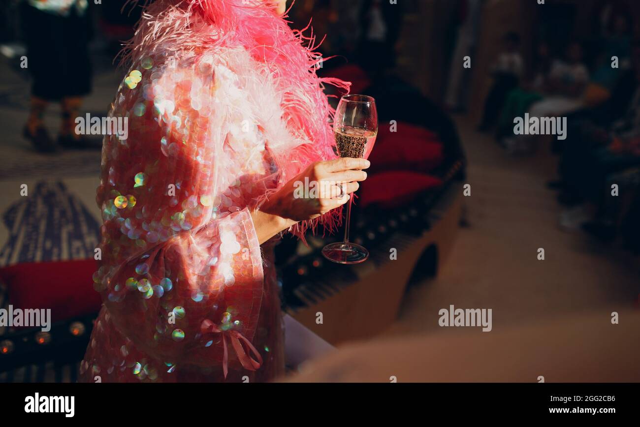 Frau in einem rosa Kleid mit Pailletten hält ein Glas Champagner in der Hand, das von der Sonne beleuchtet wird. Stockfoto