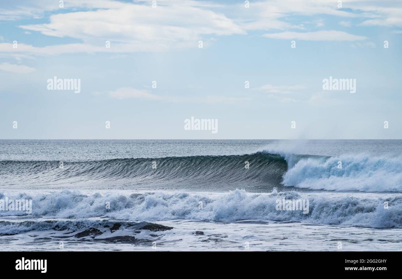 Perfekte Welle bricht direkt am Meer mit Offshore-Windbedingungen, mit blauem Himmel und Wolken Stockfoto