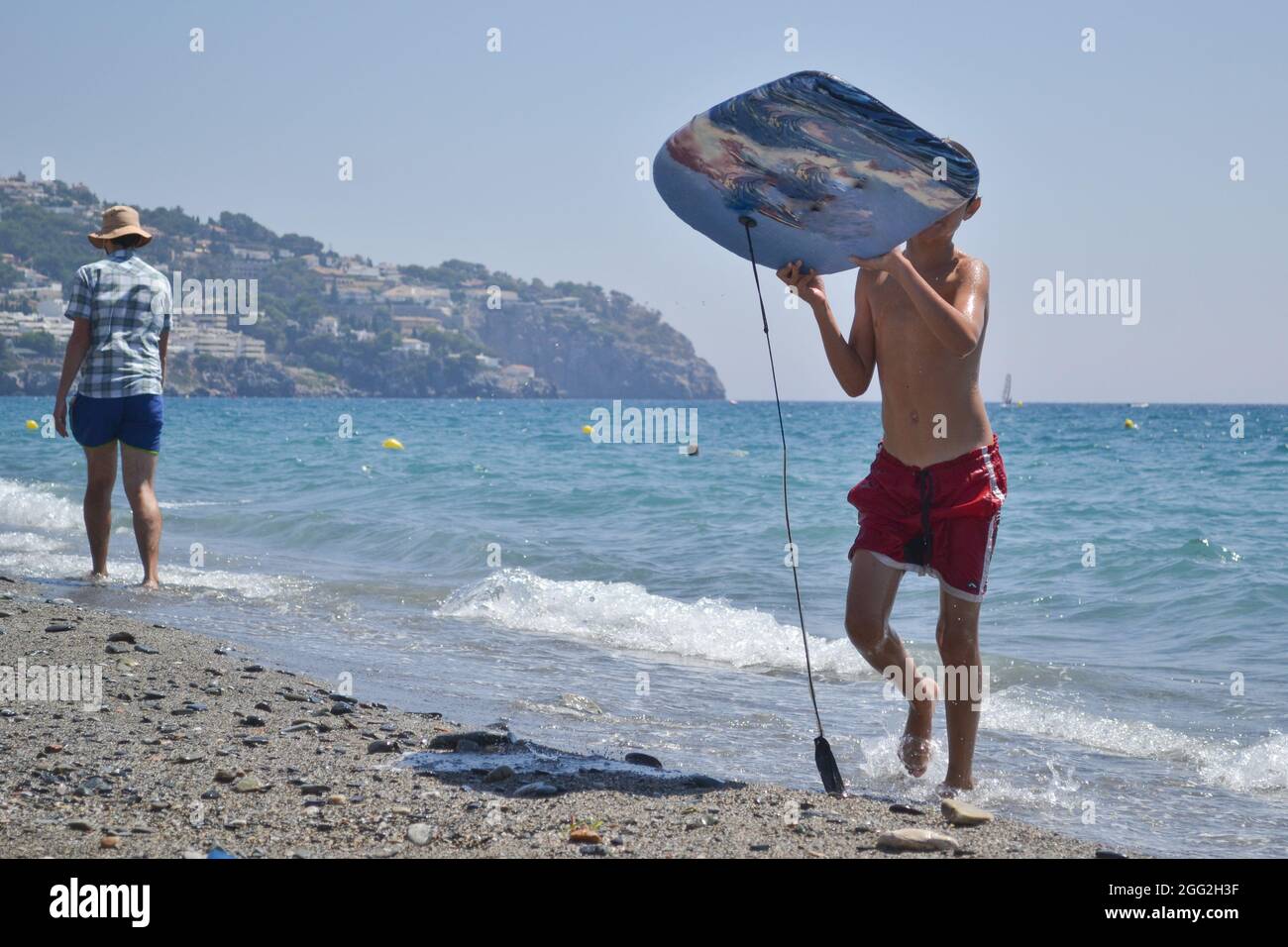 Junge, der mit Surfbrett, umgeben von anderen Badegästen, aus dem Meer kommt und Surfbretter mit Segeln im Meer Stockfoto