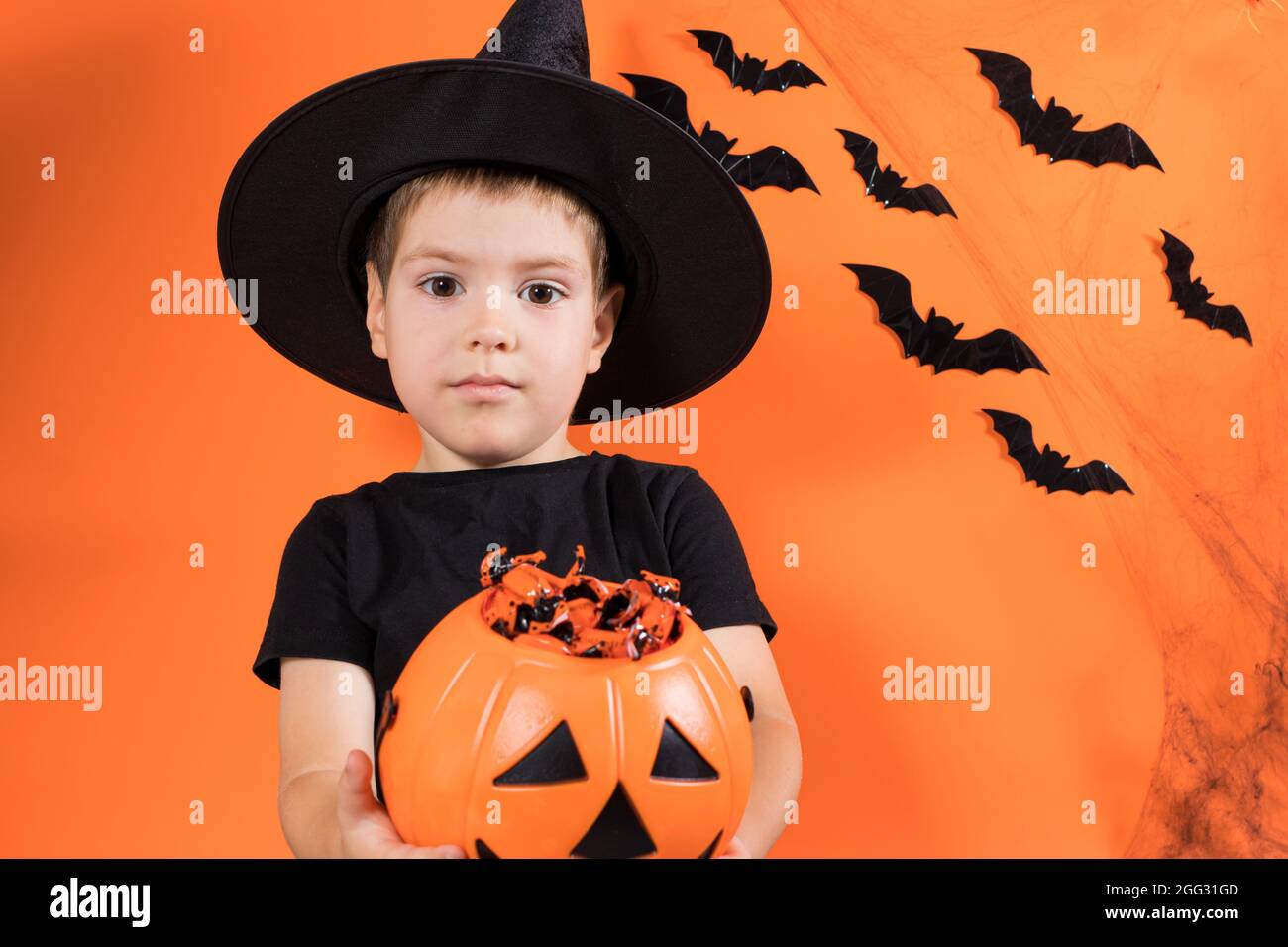 Halloween Kind. Ein Vorschuljunge in einem Hexenkostüm hält einen Kürbis mit Süßigkeiten auf einem orangen Hintergrund. Rabatte und Verkäufe für den Urlaub Stockfoto