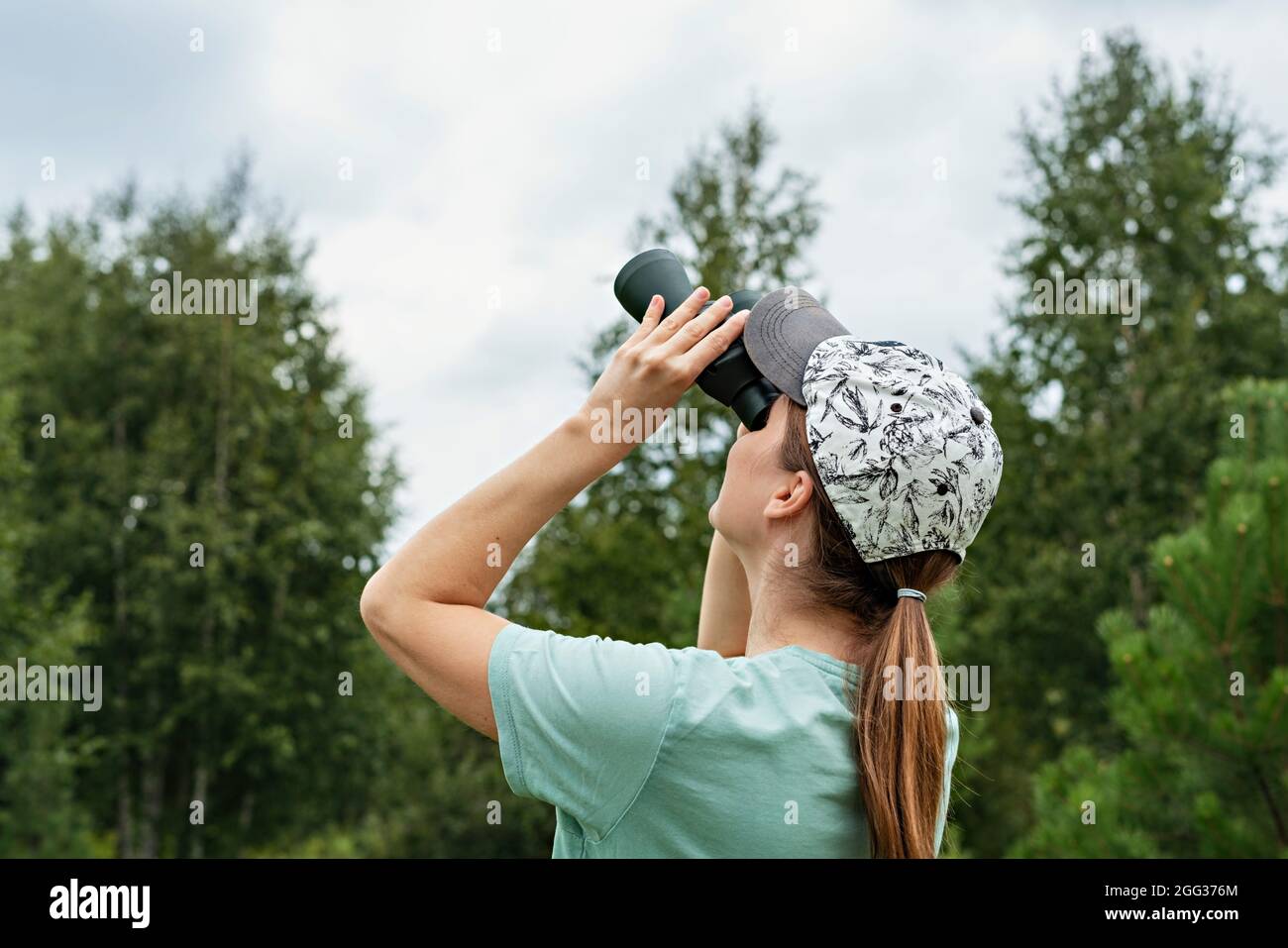 Junge blonde Frau Vogelbeobachter in Kappe und blau Blick durch das Fernglas auf den bewölkten Himmel im Sommer Wald ornithologische Forschung Vogelbeobachtung, Zoolog Stockfoto