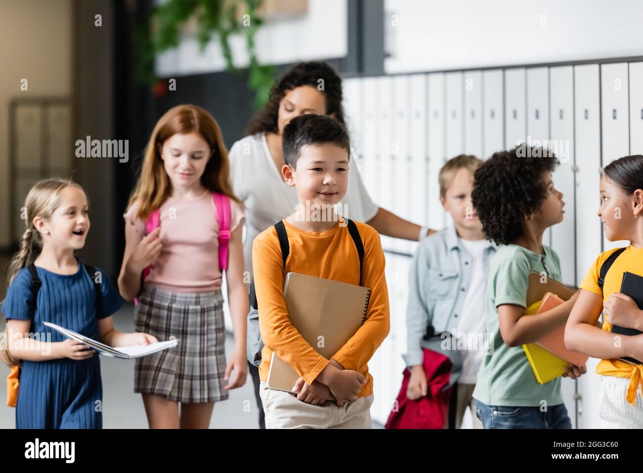 Multiethnische Schüler, die entlang des Schulkorridors in der Nähe einer verschwommenen afroamerikanischen Lehrerin gehen Stockfoto