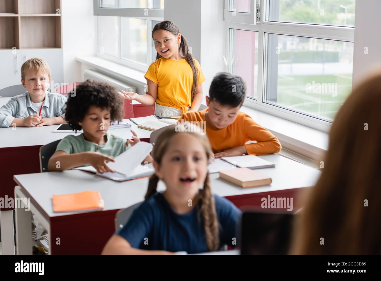 Positive asiatische Mädchen gestikulieren, während in der Nähe von multiethnischen Schülern im Klassenzimmer sprechen Stockfoto