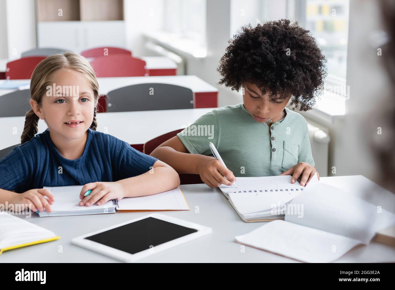 afroamerikanischer Junge, der in einem Notebook in der Nähe von Mädchen und einem digitalen Tablet mit leerem Bildschirm schreibt Stockfoto