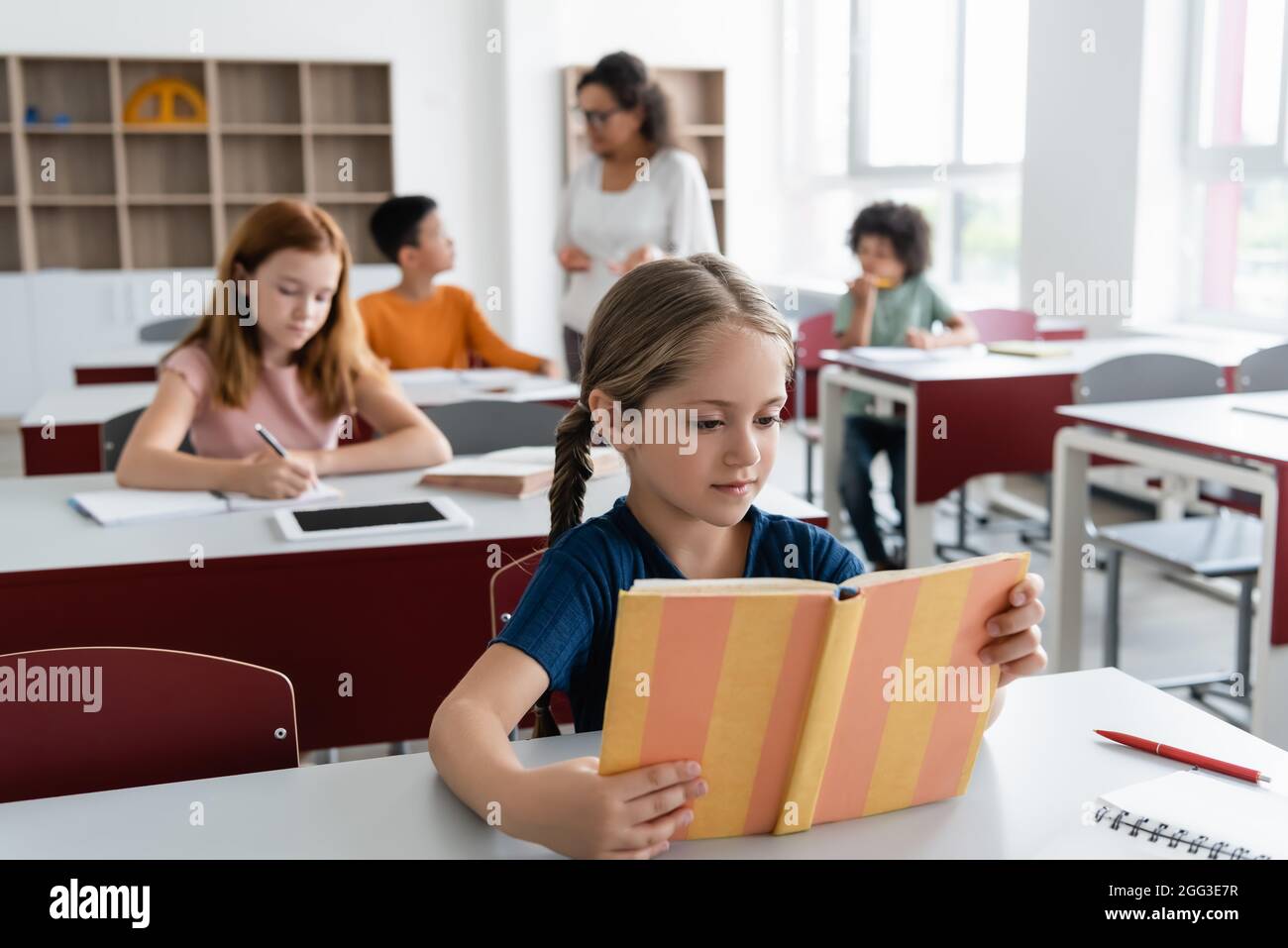 Mädchen liest Buch in der Nähe von multiethnischen Klassenkameraden und afroamerikanische Lehrerin auf verschwommenem Hintergrund Stockfoto