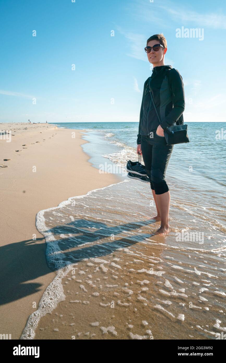 Vertikale Aufnahme einer Frau in dunkler Outdoor-Kleidung mit Schuhen in der rechten Hand, barfuß am Strand stehend, mit Gezeiten, die ihre Füße waschen. Wunderschön Stockfoto