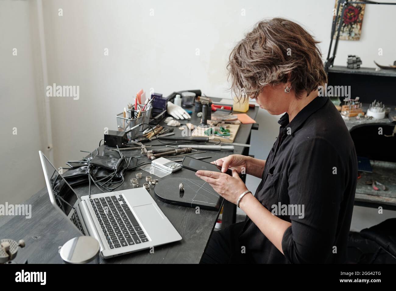 Beschäftigte Juwelierin, die in der eigenen Werkstatt sitzt und Fertigprodukte auf Marmorsteinoberflächen fotografiert Stockfoto