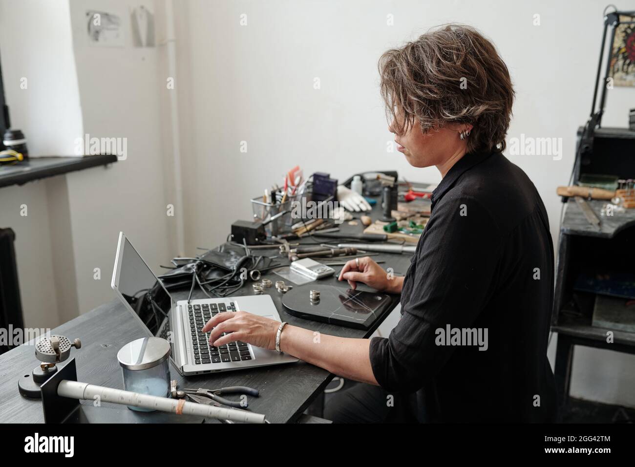 Juwelier mittleren Alters, der mit Werkzeugen am Schreibtisch sitzt und mit einem Laptop Inhalte für den Online-Juwelierladen erstellt Stockfoto