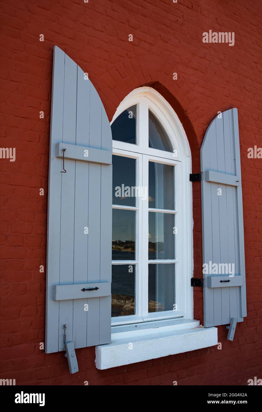 Das gewölbte Fenster mit den offenen Fensterläden auf der roten Ziegelwand des Hauses. Straßenansicht, Reisefoto, selektiver Fokus, niemand. Stockfoto