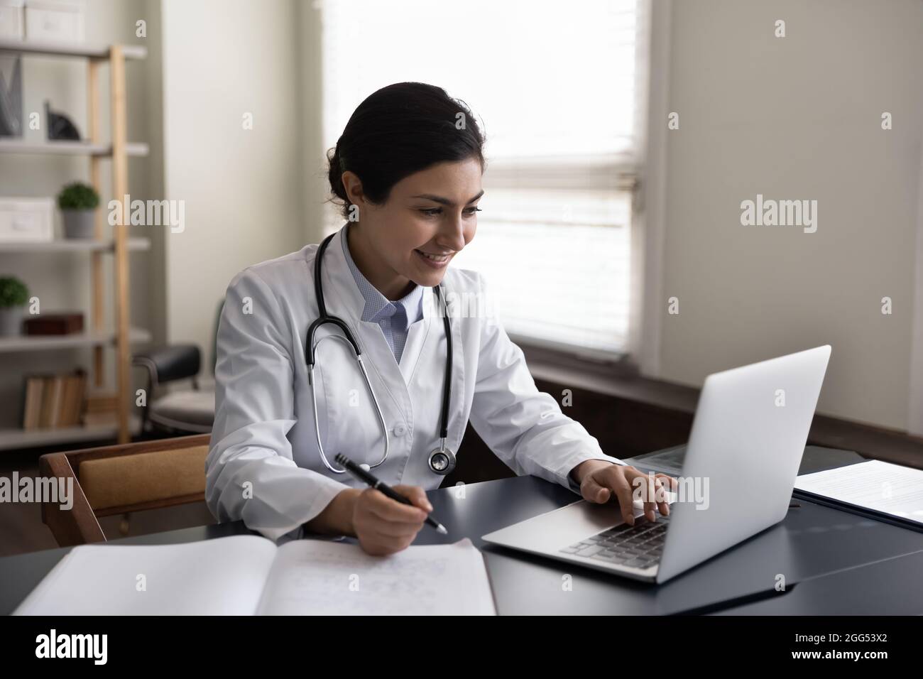 Glückliche junge indische Ärztin, die in einem modernen Klinikbüro arbeitet. Stockfoto