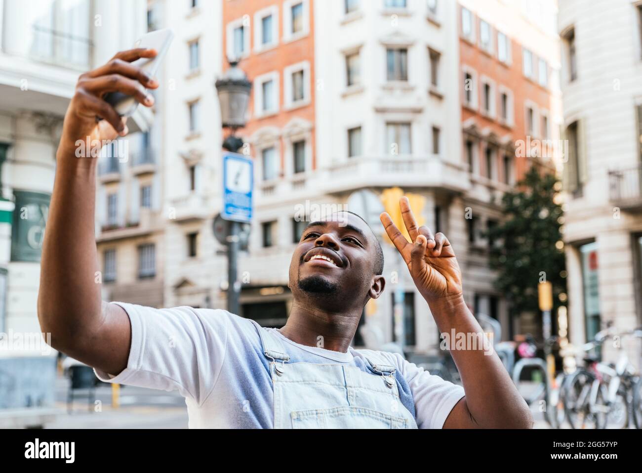 afrikanischer attraktiver Mann, der seinen Arm mit seinem Telefon hob, um ein Selfie zu machen. Er lächelt zur Kamera. Die Straßen der Stadt Barcelona im Hintergrund Stockfoto