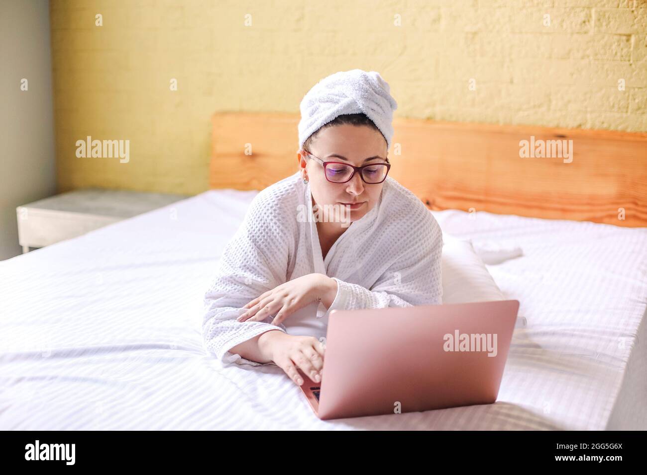 Glückliche Frau in einer Brille wartet und hat einen Videoanruf auf dem Laptop, der auf dem Bett liegt. „Work from Home“-Konzept Stockfoto