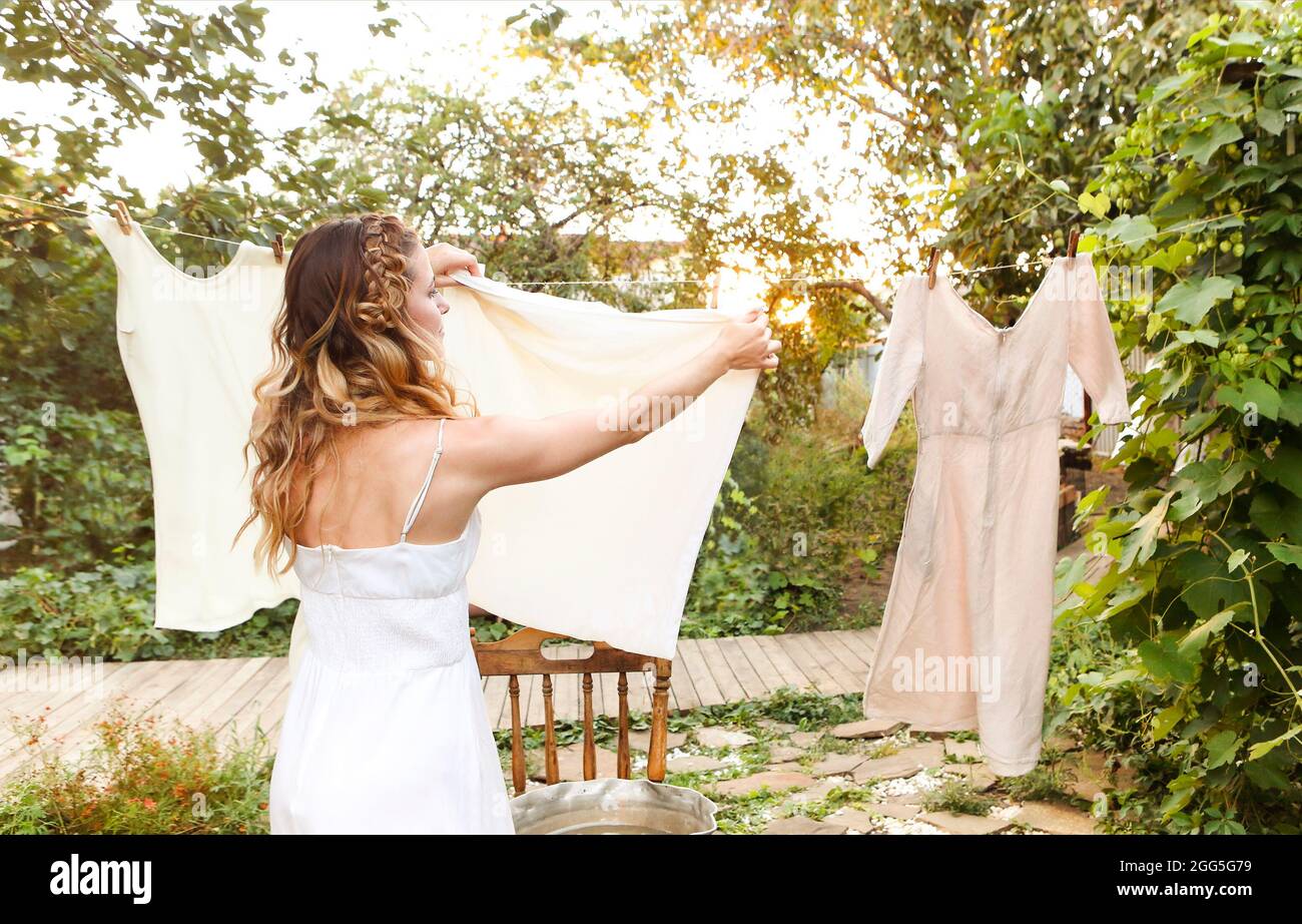 Junge Frau hängend Wäsche im Freien. Nettes Mädchen im Kleid waschen weiße Kleidung in Metall-Waschbecken im Hinterhof, hängen Wäsche auf Wäscheleine und verlassen Stockfoto