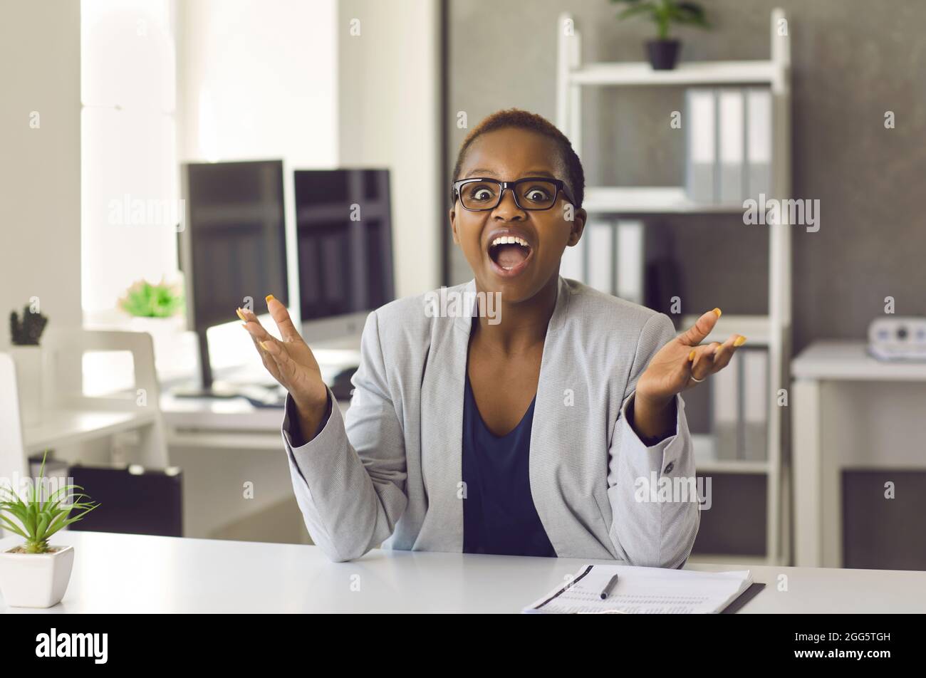 Glücklich überrascht schwarze Frau sitzt am Schreibtisch, Blick auf die Kamera und applaudiert Stockfoto