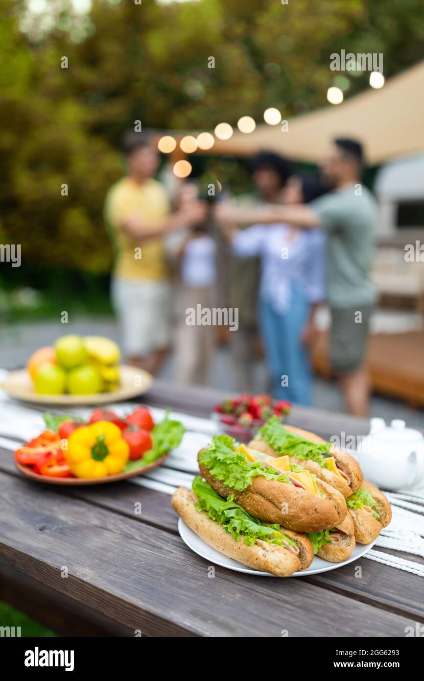 Sandwiches und Gemüse, Picknick-Speisen auf dem Tisch im Freien, Gruppe junger Freunde, die mit Bier auf dem Hintergrund toasten Stockfoto
