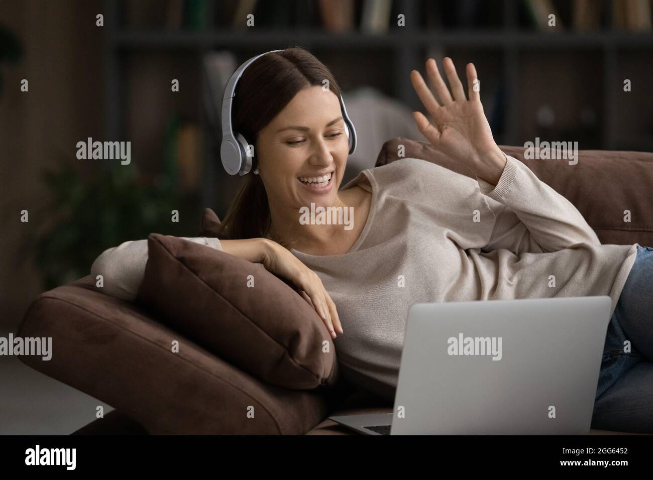 Lächelnde Frau in Kopfhörern, die mit dem Laptop auf der Couch, der Hand winkend, Stockfoto