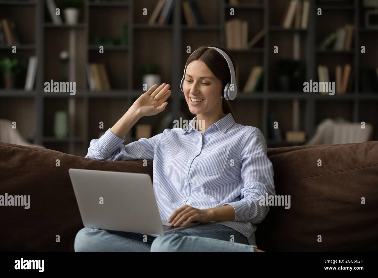 Lächelnde Frau mit Kopfhörern, die mit der Hand die Webcam anwinkt und einen Laptop benutzt Stockfoto