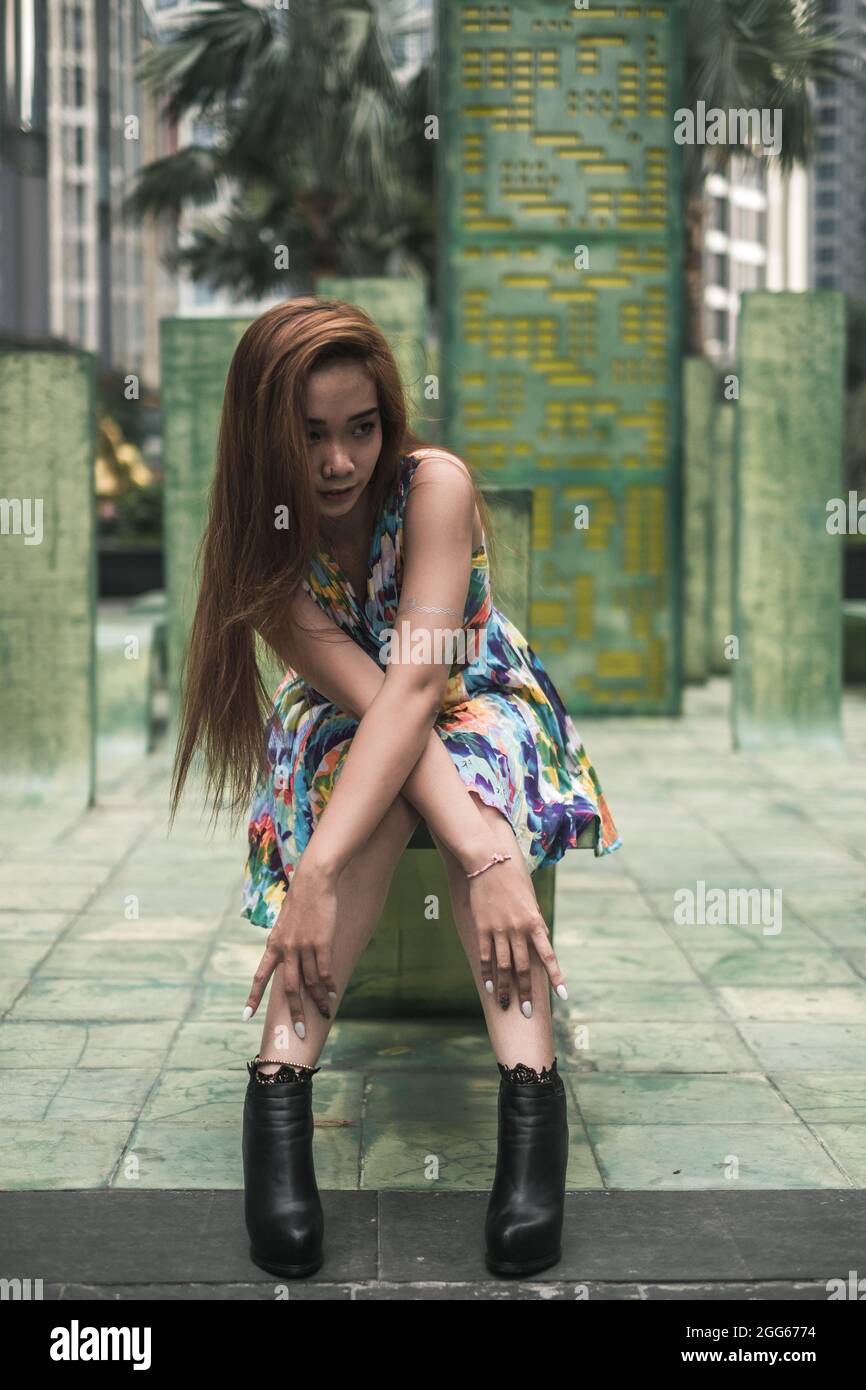 Schöne asiatische Frau in lässigen bunten Kleid sitzt im Freien in der Nähe von modernen Gebäuden. Städtisches Konzept. Langes Haar. Schöne Haltung. Hochwertige Fotos Stockfoto