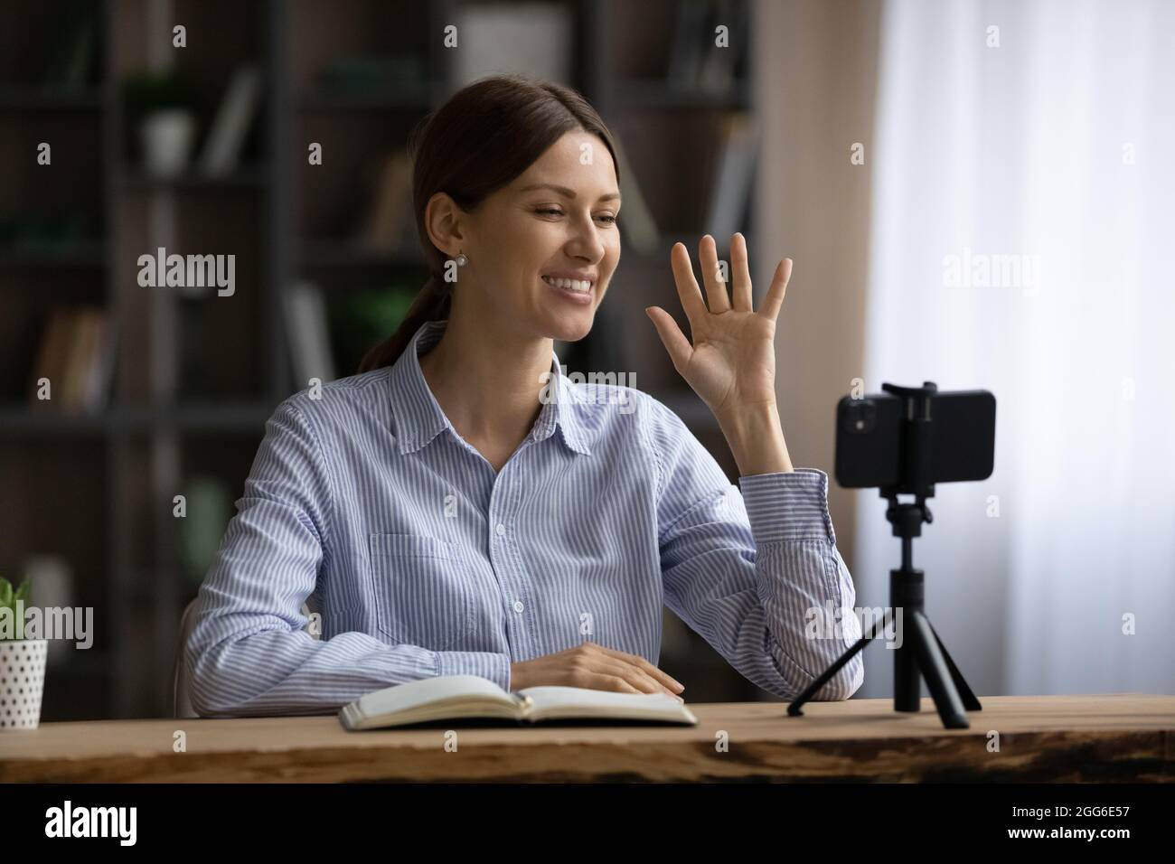 Lächelnde Frau winkt mit der Hand, nutzt das Smartphone auf dem Stativ und nimmt Videos auf Stockfoto