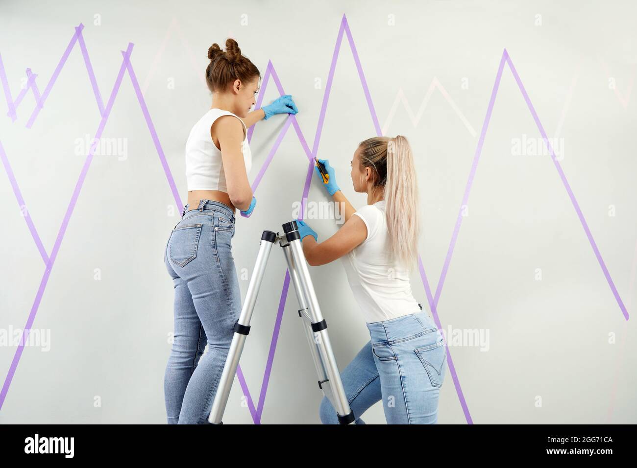 Zwei junge Frauen, die die Wand für das Malen vorbereiten, stehen auf einer  Leiter und tragen Klebeband an der Wand auf. DIY-Projekt Stockfotografie -  Alamy