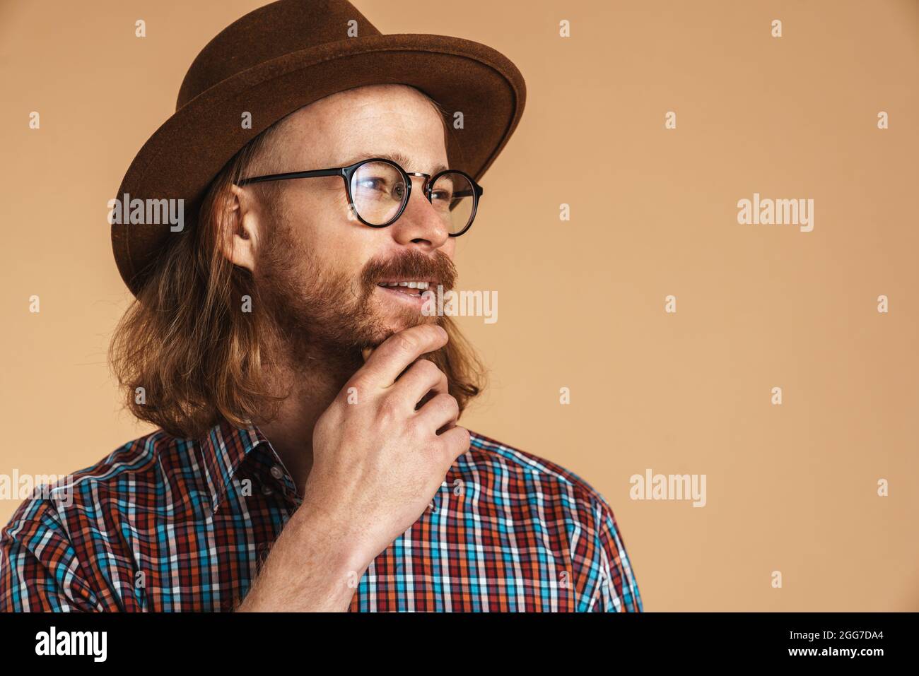 Lächelnder Ingwermann mit Brille und Hut, der isoliert auf beigen Hintergrund zur Seite schaut Stockfoto